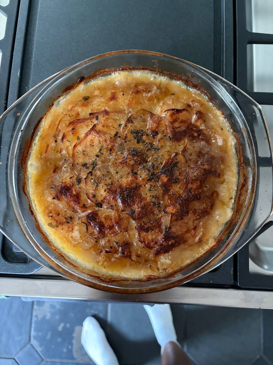 Overhead view of a round glass baking dish of boulangère potatoes fresh from the oven, showing a deeply golden and caramelised top layer of thinly sliced potatoes scattered with dark herbs, sitting on a gas hob with bubbling juices around the edges.