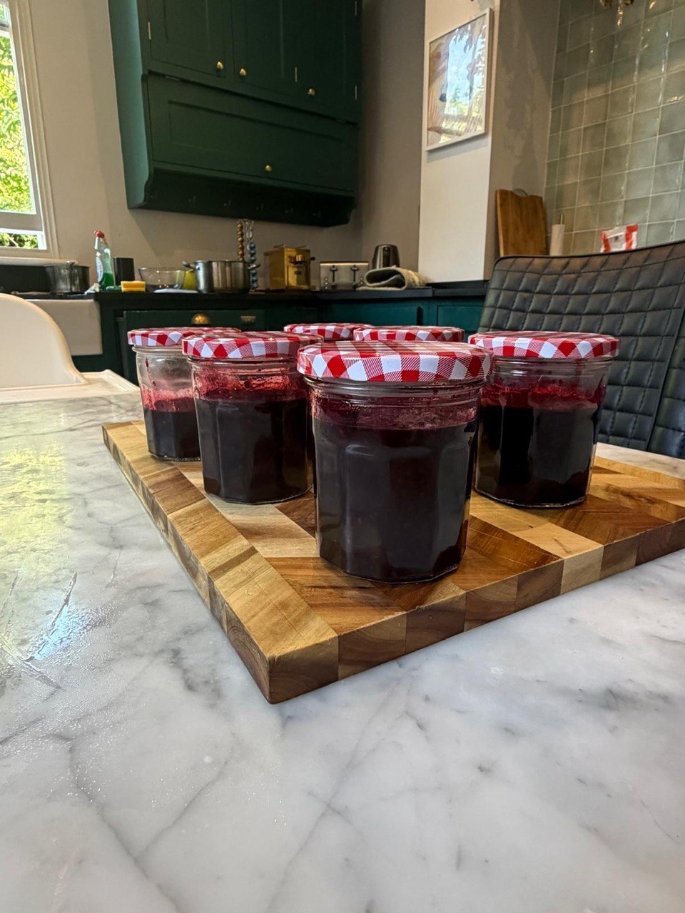 Five glass jars filled with deep purple-red blackberry jam, sealed with red and white gingham lids, arranged on a wooden chopping board on a marble kitchen counter, with dark green cabinetry visible in the background.