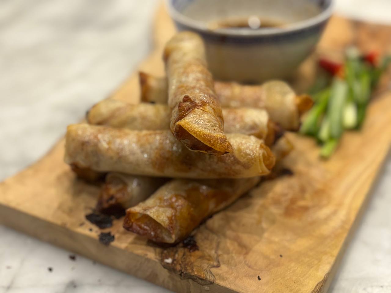 A pile of golden, crispy air-fried chicken spring rolls stacked on a wooden board, with sliced spring onions and red chilli on the side and a small bowl of dipping sauce in the background