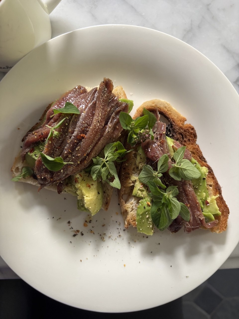 Overhead view of two slices of golden toasted sourdough on a white plate, topped with smashed avocado, layered anchovy fillets, dried chilli flakes, black pepper and fresh green herb leaves, with a white mug visible in the top corner on a marble surface