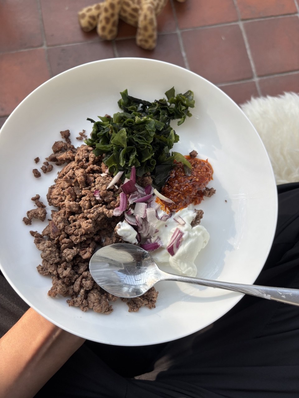 Overhead view of a white bowl held on a lap, filled with browned lean beef mince, rehydrated wakame seaweed, a dollop of white yogurt topped with red harissa paste, and diced raw red onion, with a silver spoon resting in the bowl