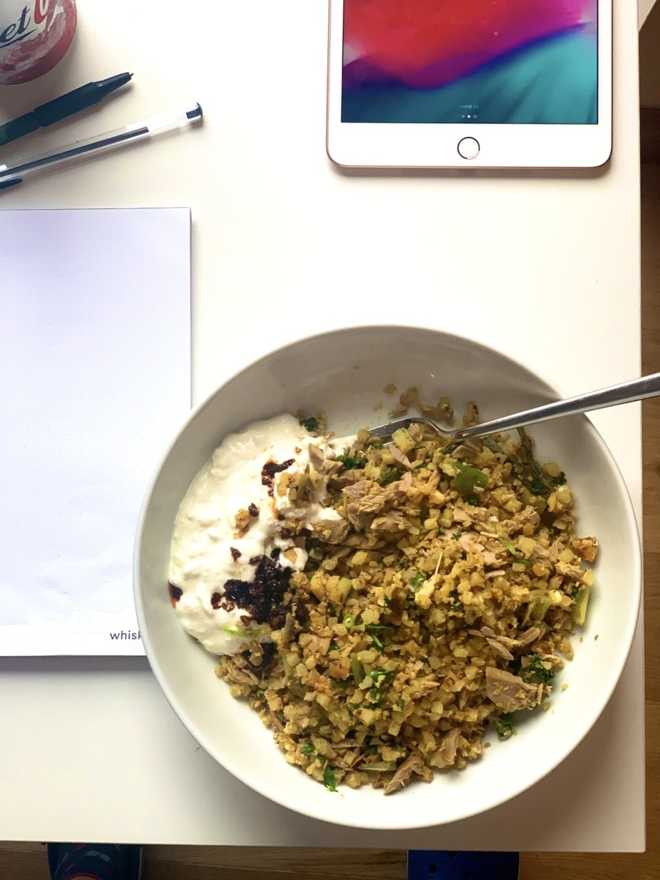 Overhead view of a white wide-rimmed bowl containing golden spiced cauliflower fried rice with flaked tuna and chopped parsley, with a dollop of white yoghurt drizzled with chilli oil on one side and a fork resting in the bowl. An iPad, pens and a notepad are visible on the white desk around it.
