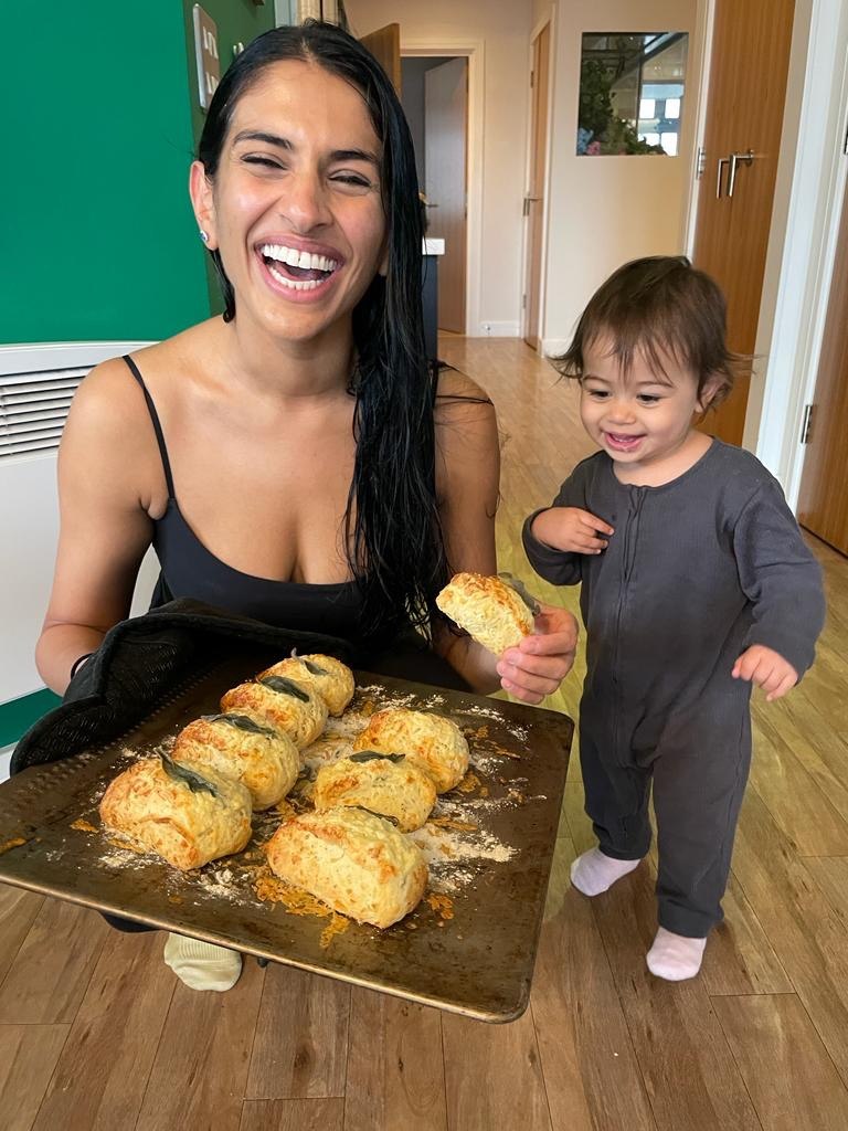 A laughing woman holds a dark baking tray loaded with golden sage and cheddar scones toward the camera. A smiling toddler in a grey sleepsuit stands beside her, already holding one scone in their hand.