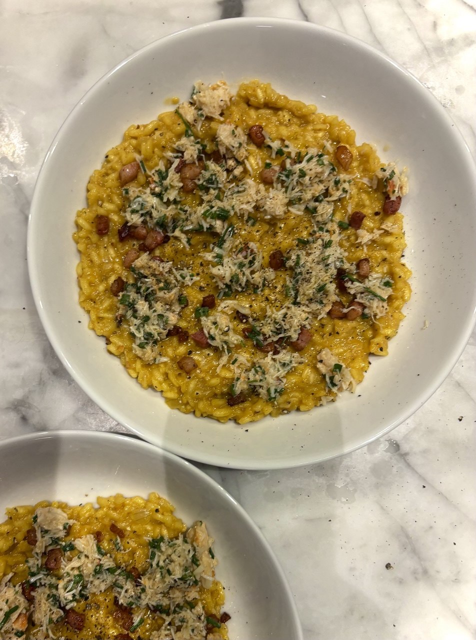Overhead view of two white bowls on a marble surface, each filled with golden saffron risotto topped with flaked white crab meat, crispy pancetta cubes, finely snipped chives and a dusting of grated Parmesan