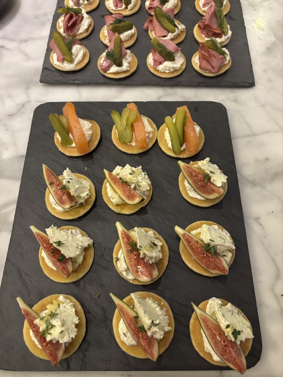 Two slate serving boards covered with small round blinis. The front board shows two rows of fig and herb cream cheese blinis with fresh fig slices and thyme leaves, and one row of smoked salmon and cornichon blinis. The back board shows blinis topped with folded slices of cured meat, cornichons and dill, on a white marble surface.