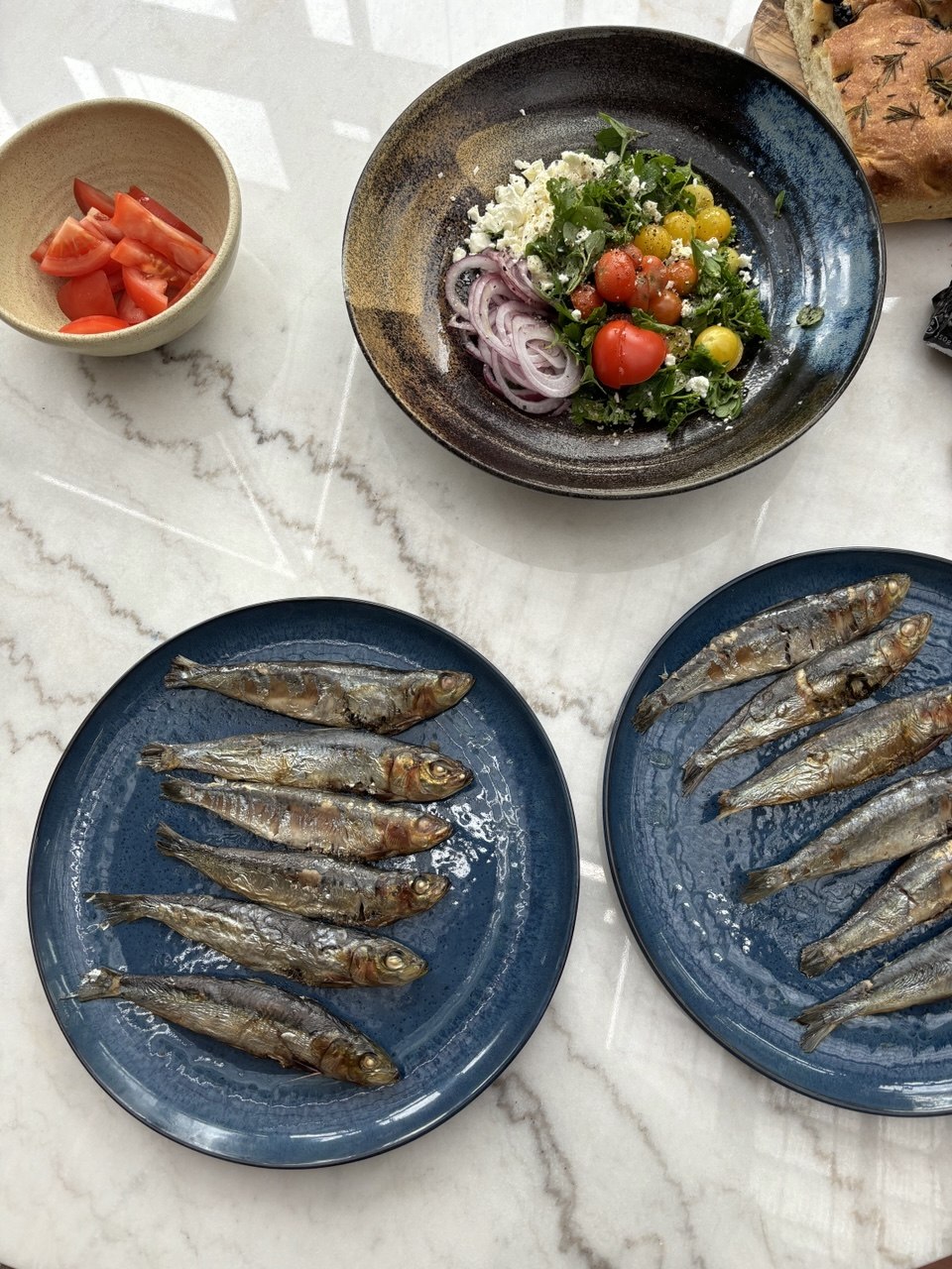 Overhead view of a marble table set with two blue plates each holding five or six whole grilled sardines, a dark ceramic bowl containing a salad of cherry tomatoes, sliced red onion, green herbs and crumbled feta, a small cream bowl of tomato wedges, and rosemary focaccia at the edge of the frame