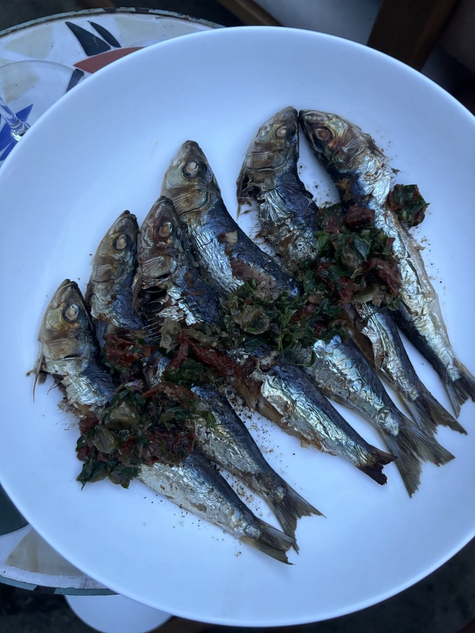 Overhead view of around ten whole grilled sardines piled in a large white bowl, topped with a rough dressing of chopped sun-dried tomatoes and fresh green herbs.