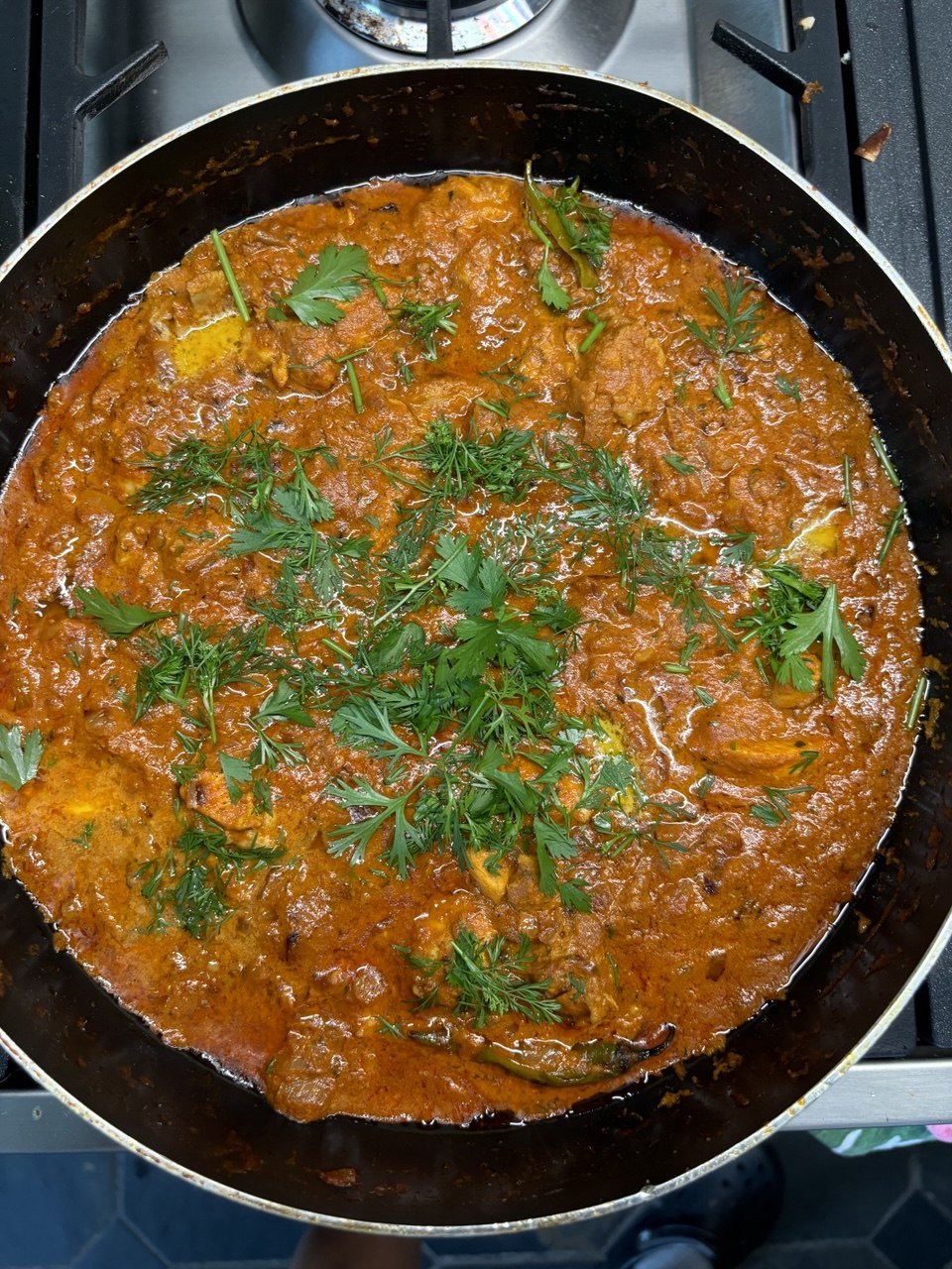 Overhead shot of a large dark karahi pan on a gas hob, filled with a vibrant deep-orange butter chicken curry garnished with fresh coriander leaves and slit green chillies.