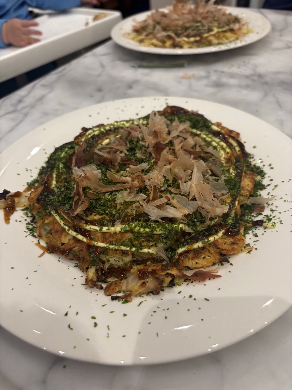 A round Japanese savoury pancake on a white plate, topped with concentric swirls of dark brown sauce and pale mayo, a heavy dusting of bright green aonori seaweed flakes, and a generous pile of thin, curling bonito flakes in the centre. A second similar plate is visible in the background on a marble surface.