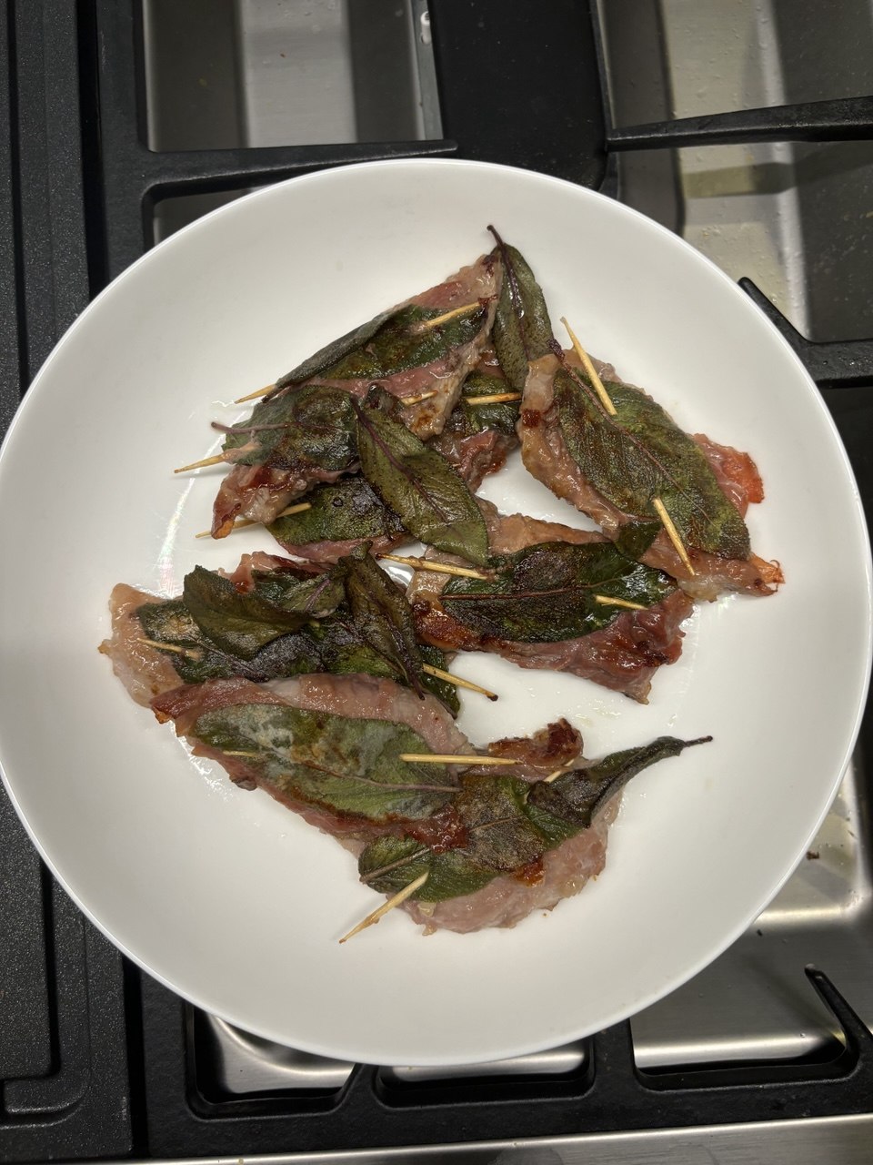 Eight cooked veal escalopes topped with crisped sage leaves and prosciutto, secured with cocktail sticks, piled on a white plate resting on a gas hob