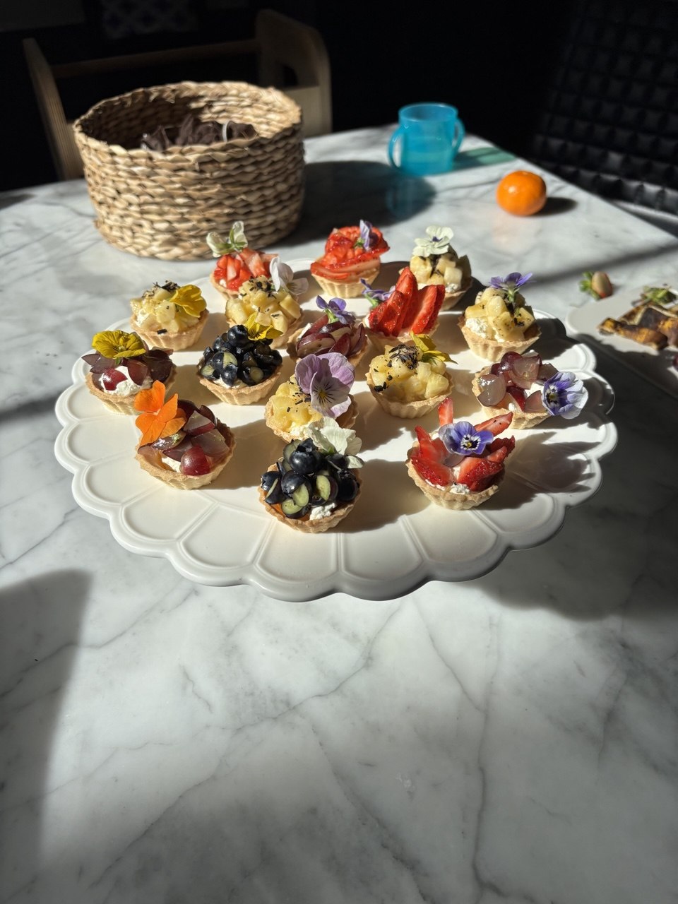 Sixteen small wafer cups arranged on a white scalloped cake stand on a marble table, each filled with whipped ricotta cream and topped with a colourful mixture of strawberries, blueberries, red grapes and pineapple, decorated with purple, yellow and orange edible flowers including pansies and nasturtiums, photographed in bright natural sunlight