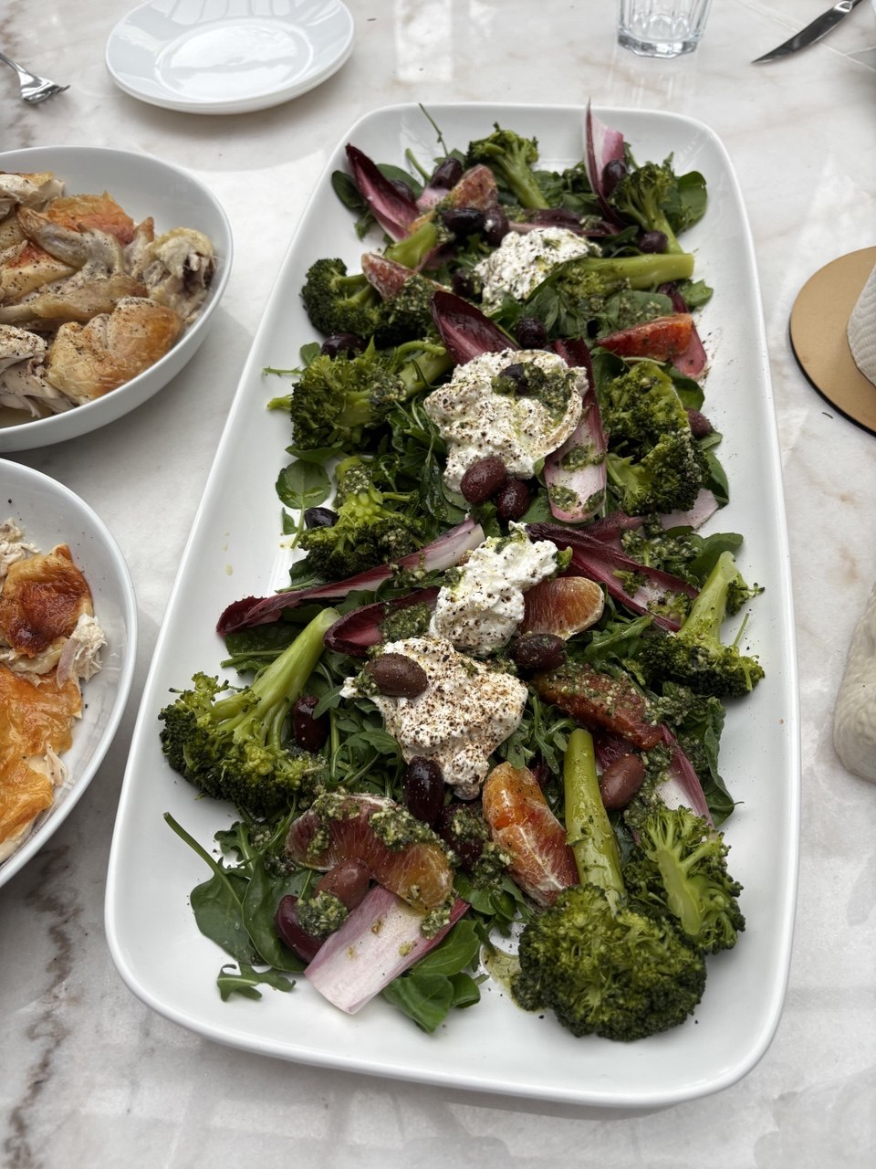 A large white rectangular platter loaded with roasted broccoli florets, torn burrata, blood orange slices, kalamata olives and radicchio leaves on a bed of rocket, drizzled with a vivid green herb oil and finished with cracked black pepper, photographed from above on a marble table alongside bowls of chicken dishes.