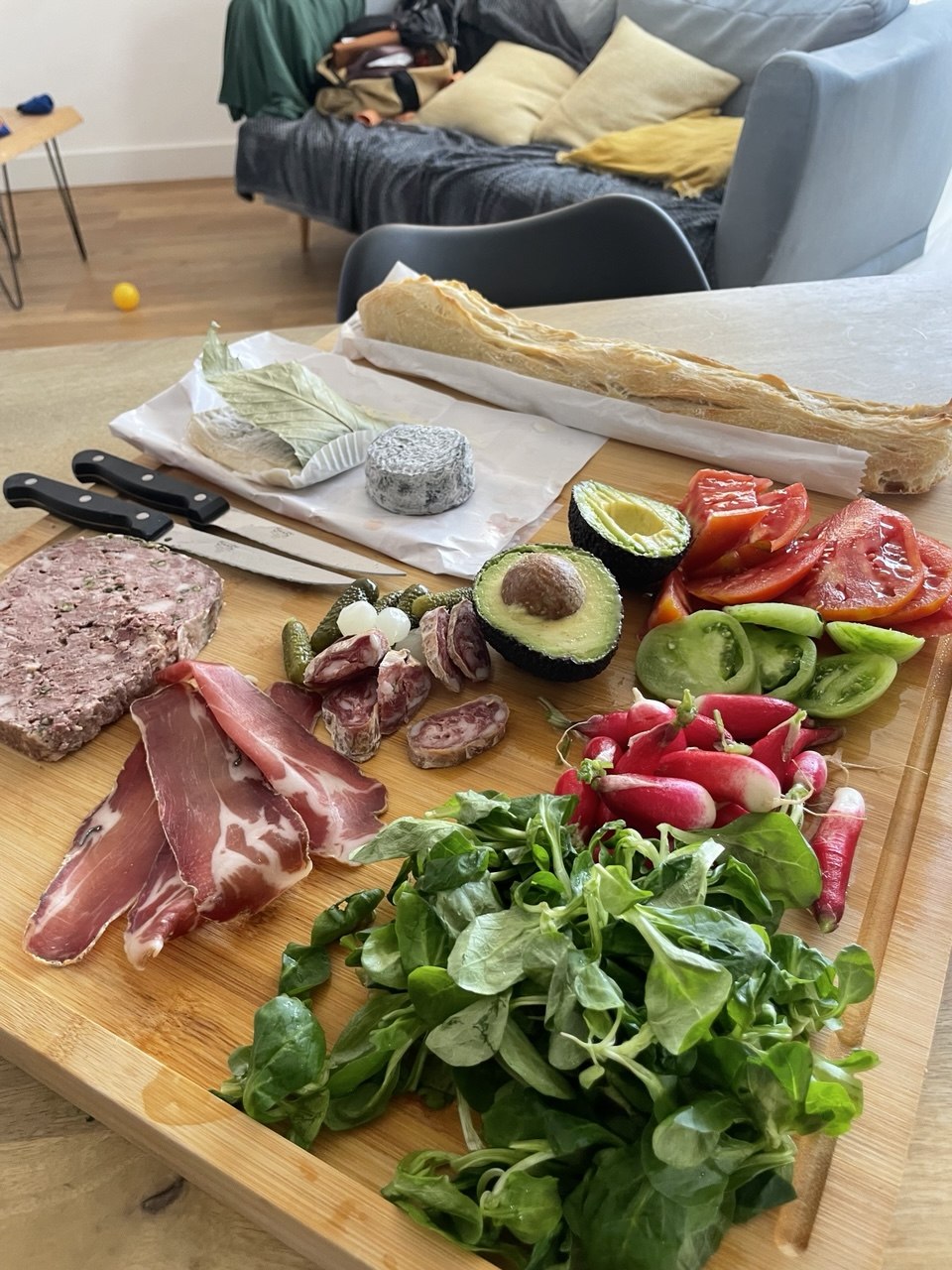 A large bamboo board set on a coffee table, loaded with sliced pâté, cured ham, salami, an ash-rinded goat's cheese round, two avocado halves, sliced red and green heirloom tomatoes, a bunch of radishes, cornichons, pickled onions, a mound of lamb's lettuce and torn sourdough baguette, with two knives resting to one side. A grey sofa with yellow cushions is visible in the background.
